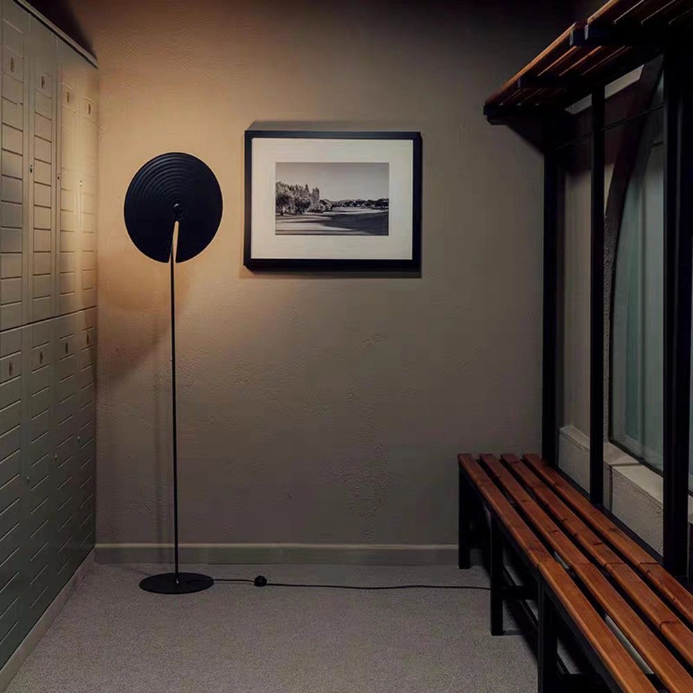 Minimalist matte black floor lamp with adjustable lighting placed in a modern interior setting, featuring a hardwood floor and neutral walls, with art and architecture elements in the background.