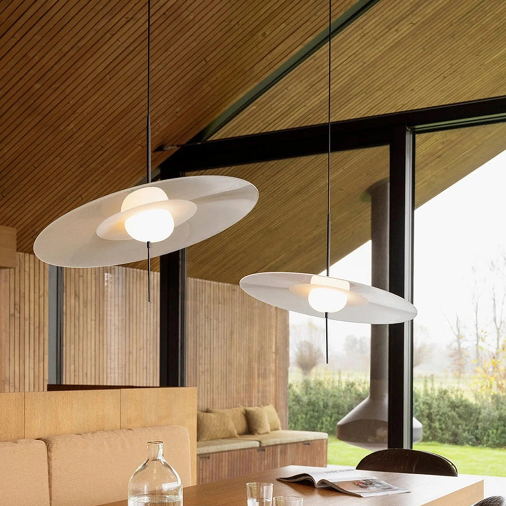 Minimalist round pendant light in white with a diameter of 15.7 inches, hanging from the ceiling in a modern interior setting. The light fixture is above a wooden table with a small potted plant and a bottle, against a simple wall backdrop.