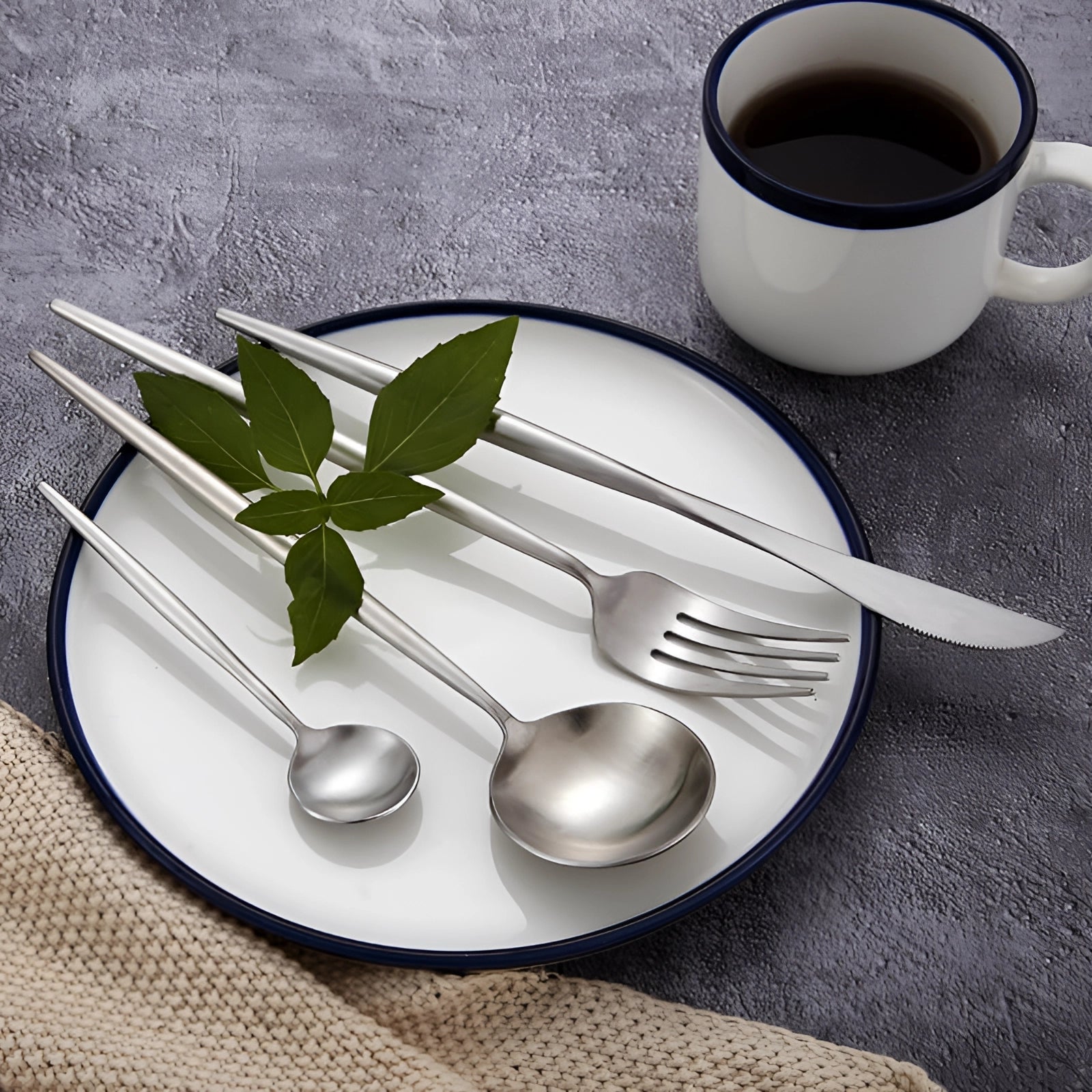 Minimalist stainless steel cutlery set displayed on a table with white dishware, including a plate and cup, showcasing an elegant and modern flatware design.
