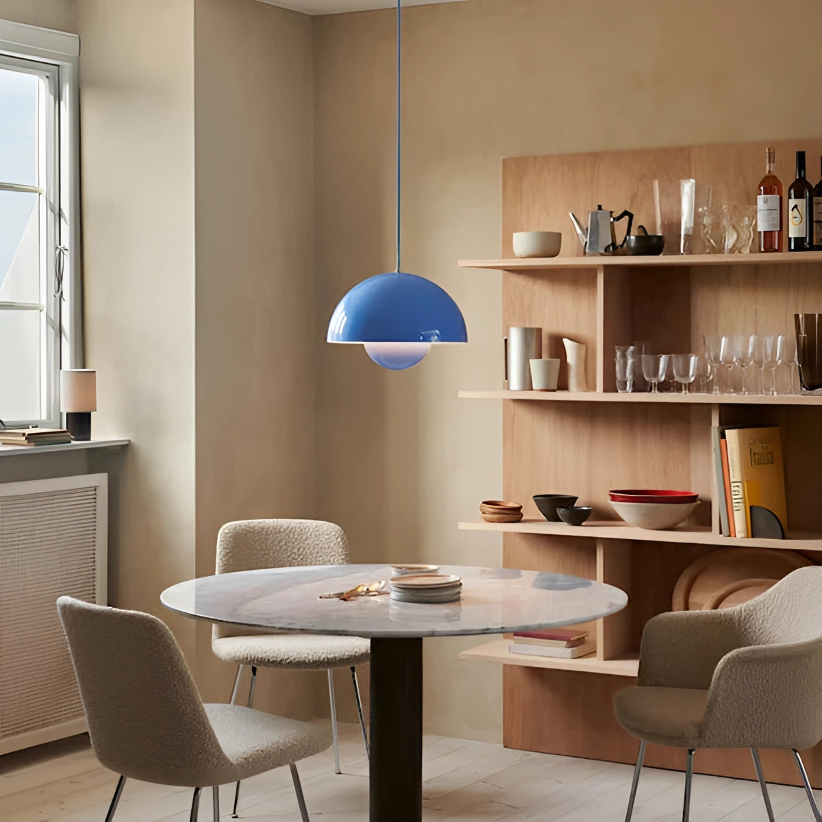 Minimalist white dome pendant light with a blue hue hanging above a dining table in a Scandinavian-style interior, featuring wooden furniture and shelving.