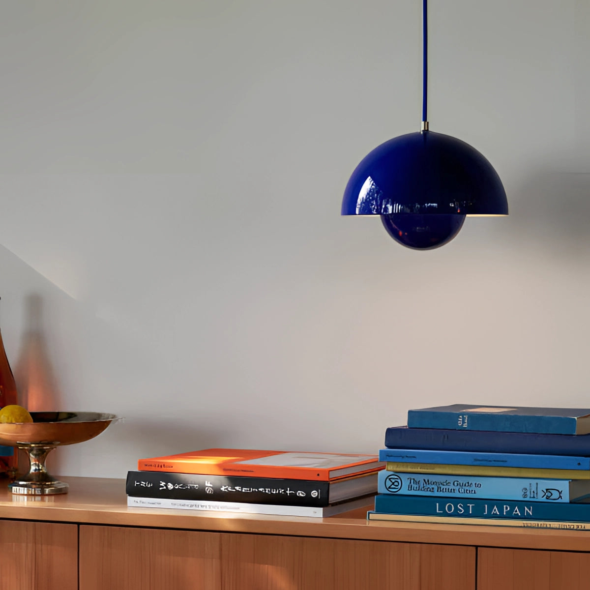 Minimalist white dome pendant light with a dark blue finish, hanging in a Scandinavian-style dining and kitchen area. The background features wooden shelving with books and decor, complementing the modern interior design theme.