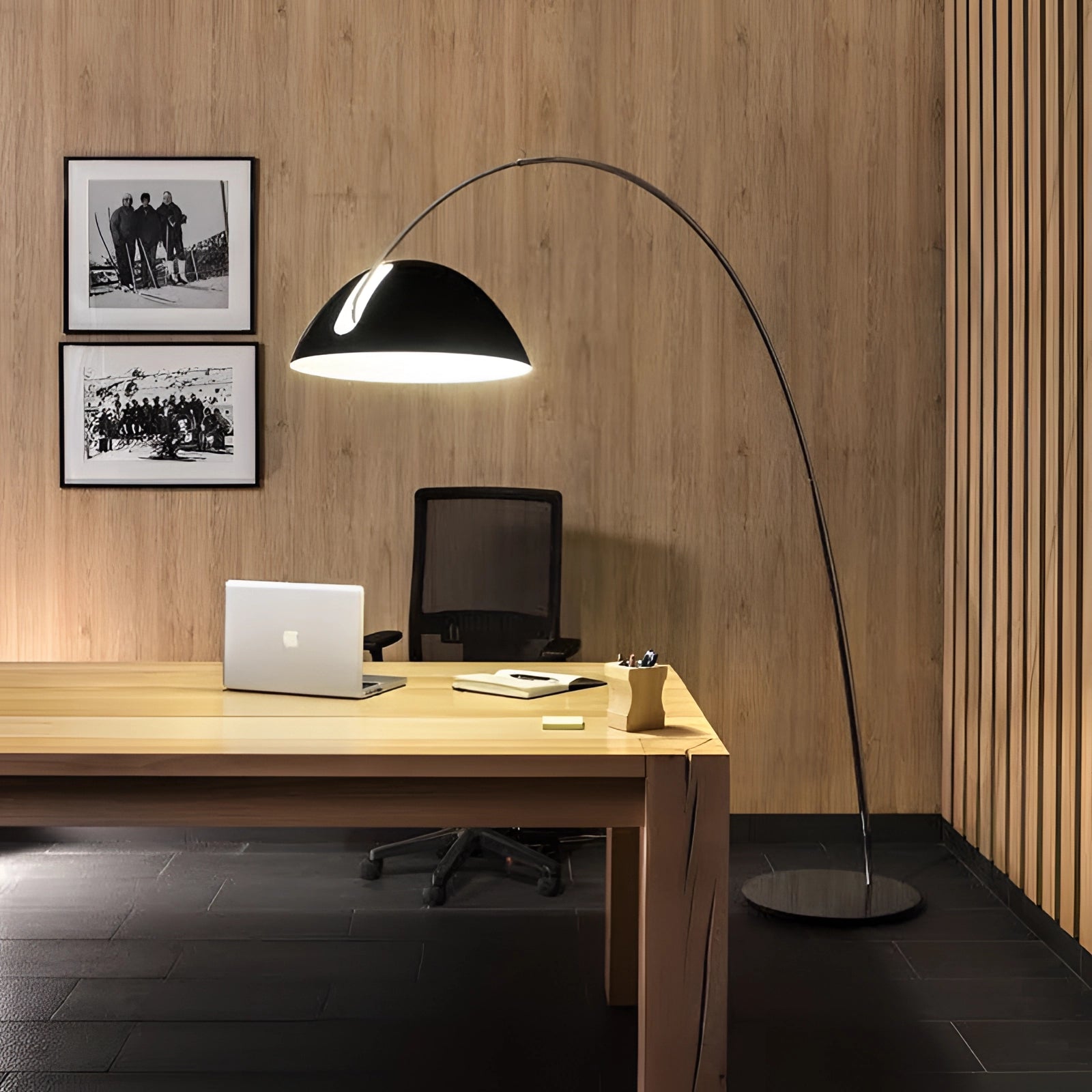 Modern black arched floor lamp with an adjustable dome reading light placed next to a wooden desk with a laptop and other office items, illustrating a stylish interior design setup.