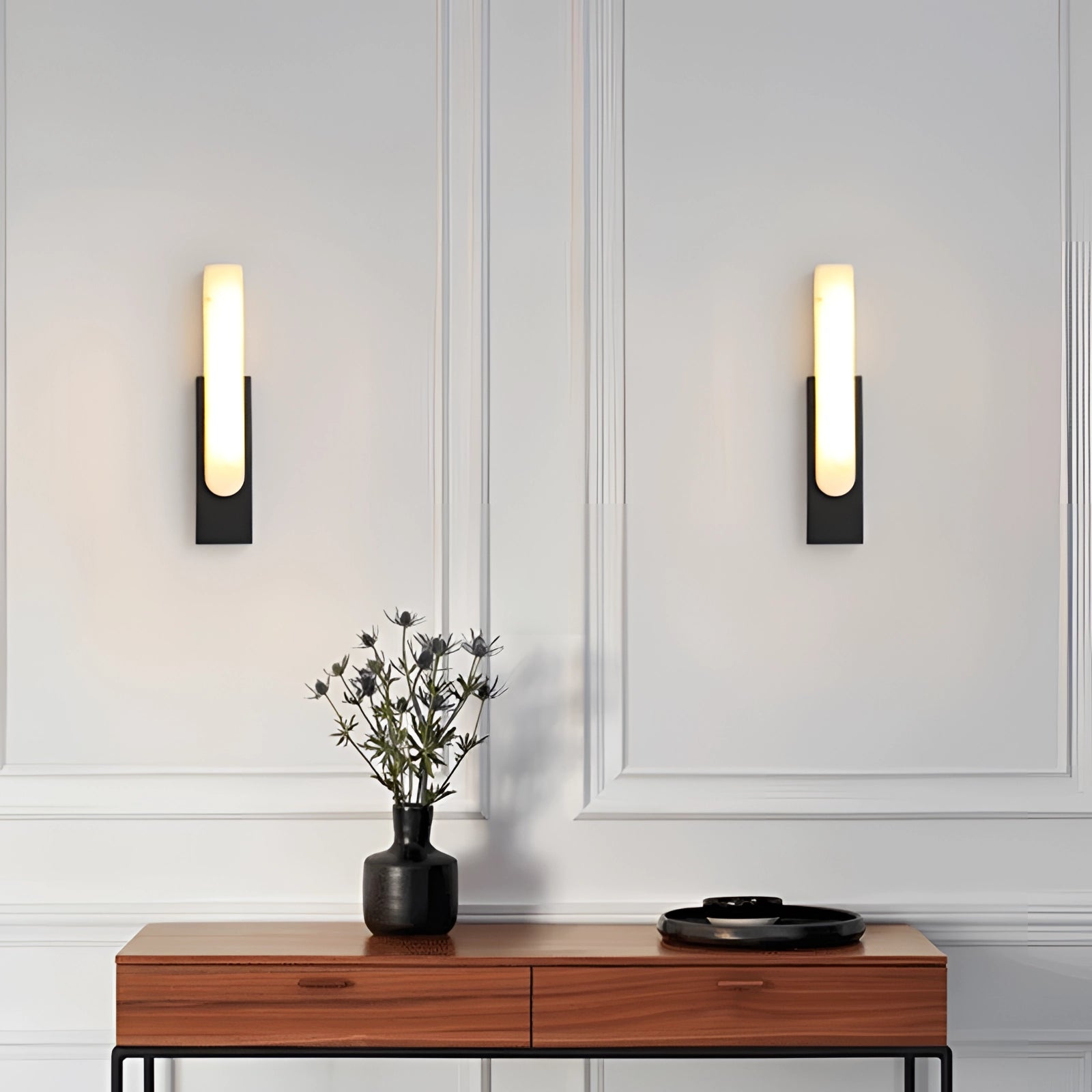 Modern black and gold LED wall sconce mounted on a wall, illuminating a minimalist indoor space with a contemporary design. The sconce is featured in a living room setting with a wooden table, a vase, and a houseplant nearby.