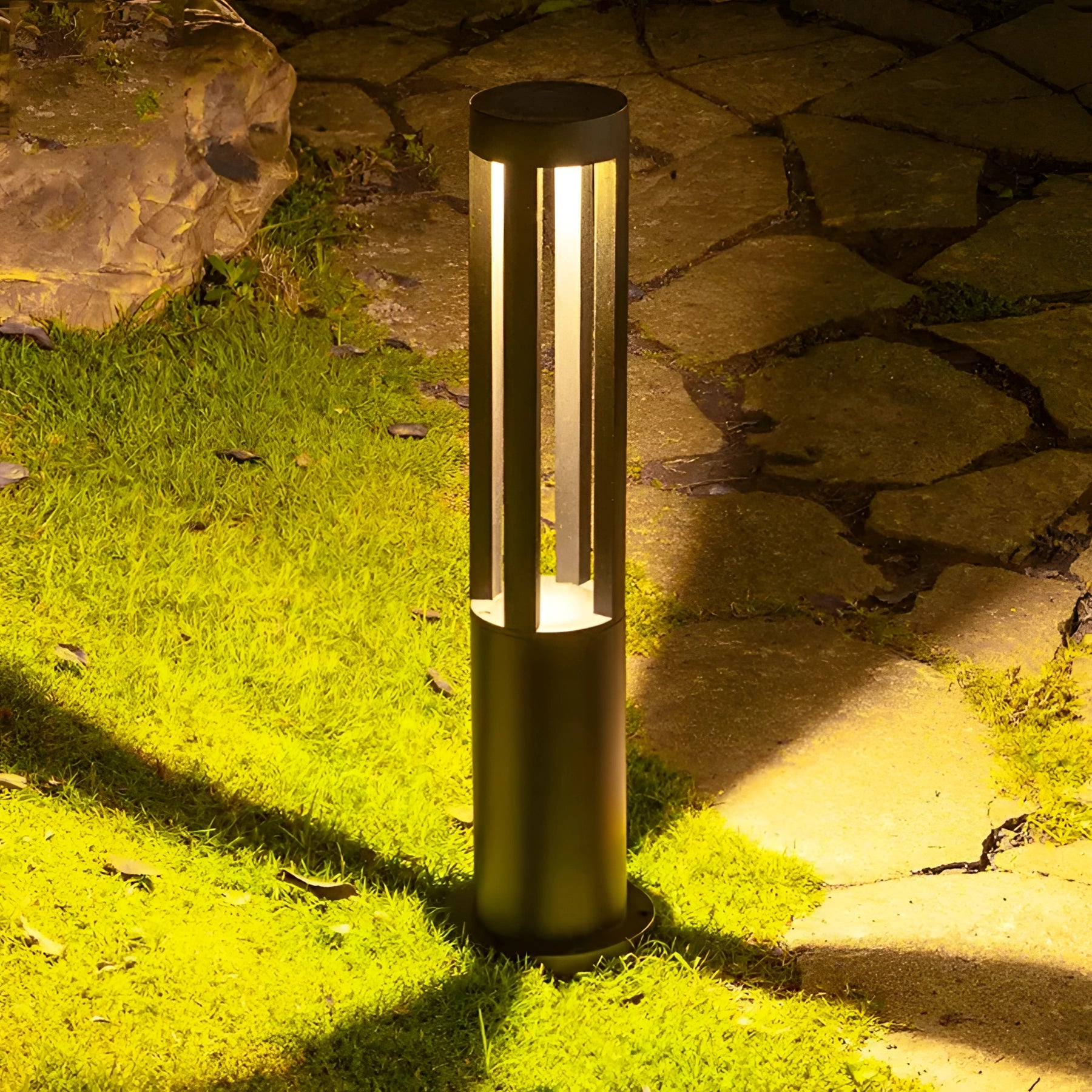Modern black LED cylinder lamp installed outdoors on a grassy area, with a sleek rectangular design and waterproof features, casting a warm yellow glow against a nearby wall.