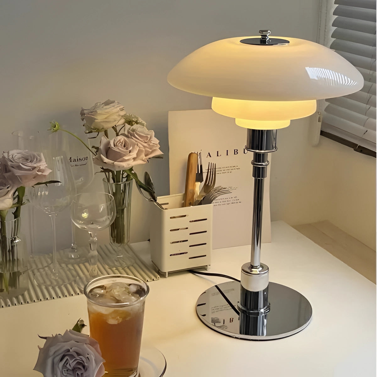 Modern black mushroom shade table lamp with adjustable height and frosted glass, placed on a table with various tableware items and a plant in the background.