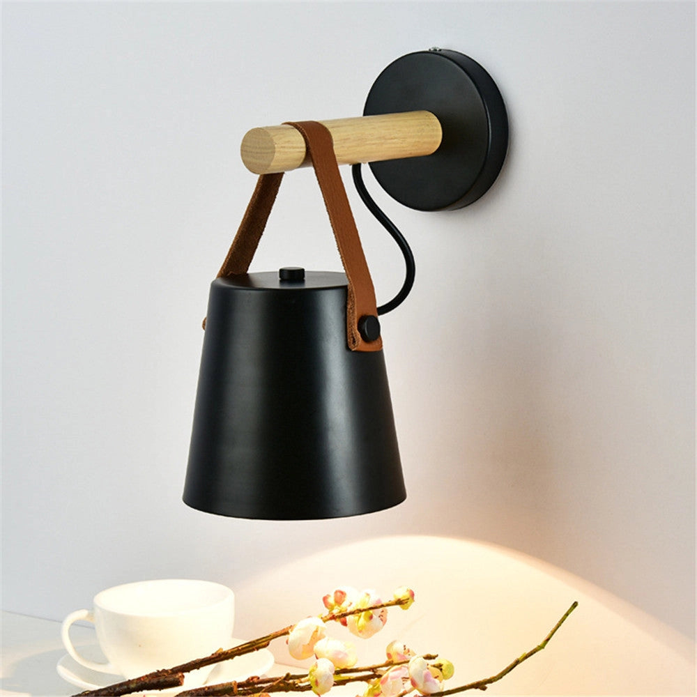 Modern black shade wall sconce with leather strap and wood accent, mounted on a wall, illuminating a room.