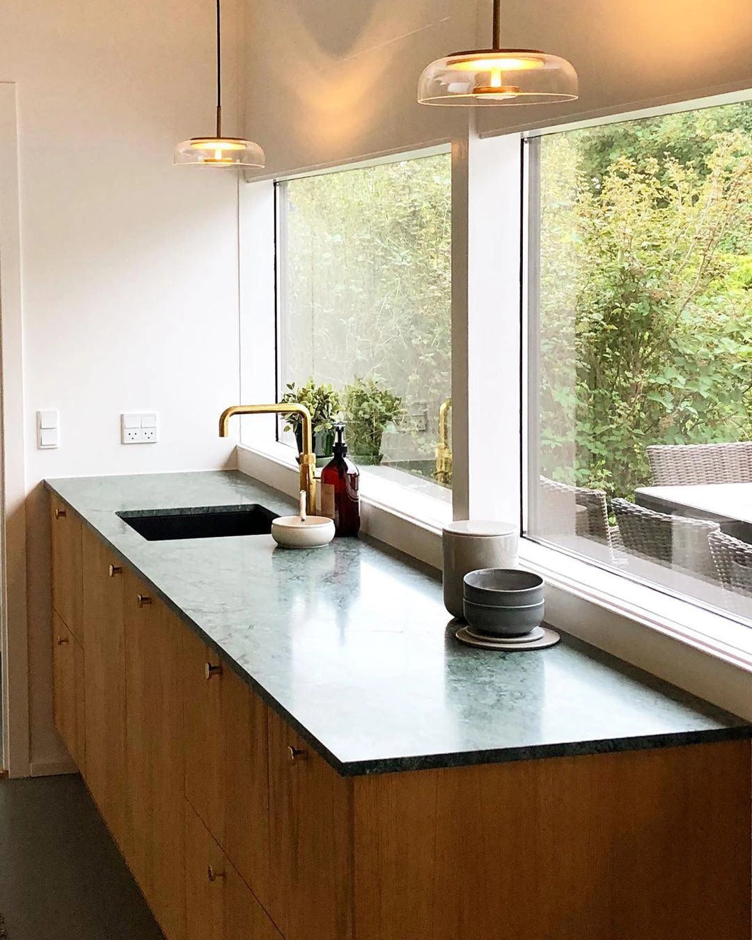 Modern brass and chrome LED pendant lights hanging above a sleek kitchen countertop with a sink, tap, and cabinetry, accented by a small plant.