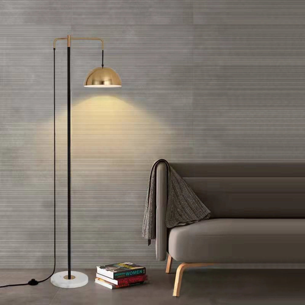 Modern brass dome shade floor lamp with adjustable gold-tone lighting sits on a wooden floor in a stylish interior space, emitting a warm white light. The setting features grey elements contributing to a comfortable and contemporary design.