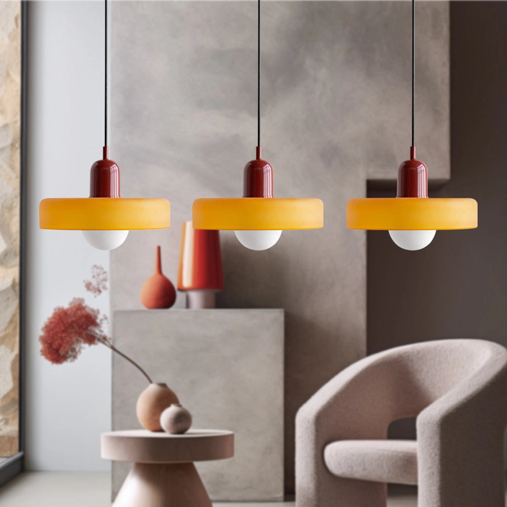 Modern brass pendant lights with three heads and adjustable red cable, featuring an orange exterior, hanging in an industrial kitchen with wooden shelving and plants.