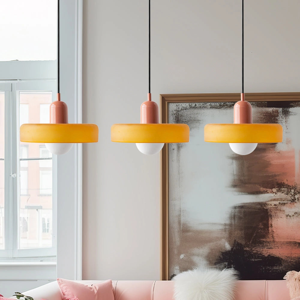 Modern brass pendant light with a red cable, hanging in an industrial kitchen setting with shelves and a large window in the background.