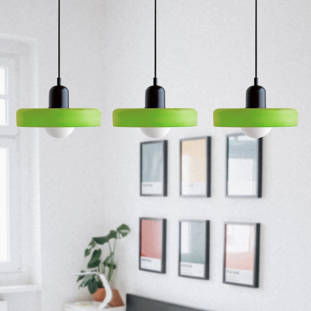 Modern industrial-style pendant lights with brass finish and red cables, featuring three matte green and black adjustable heads, hanging above a stylish kitchen area with wooden shelving and houseplants.