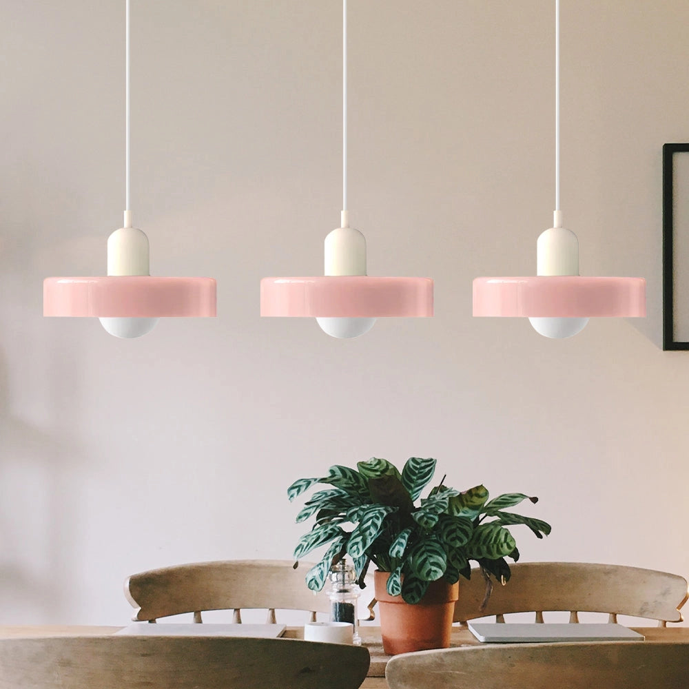 Modern brass pendant lights with adjustable red cables, featuring a pink and white design with three heads, hanging in an industrial kitchen setting with houseplants and wooden furniture.