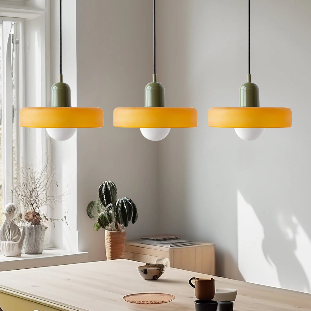 Modern brass pendant lights with three heads and red cable, featuring orange and green accents, hanging above a kitchen table with tableware and a plant in an industrial-style kitchen.