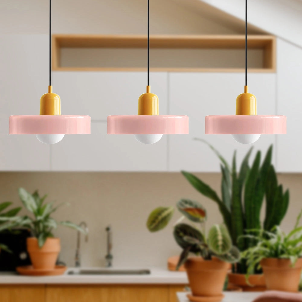 Modern brass pendant lights with red cable, featuring three heads in pink and yellow, hanging above a stylish industrial kitchen with wooden shelving and various houseplants.