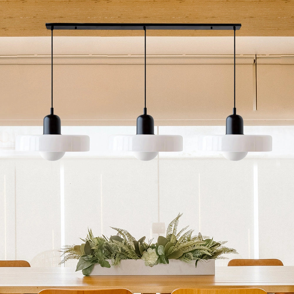 Modern brass pendant lights with a red cable, featuring a black and white design and three adjustable heads, hanging above a wooden shelf with houseplants and decorative items in an industrial kitchen.