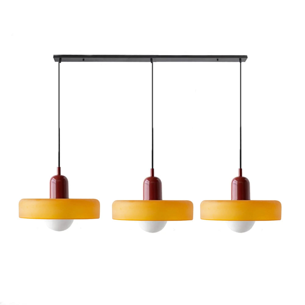 Modern brass pendant light with an adjustable red cable, hanging above a kitchen island with shelves, plants, and decorative items in the background.