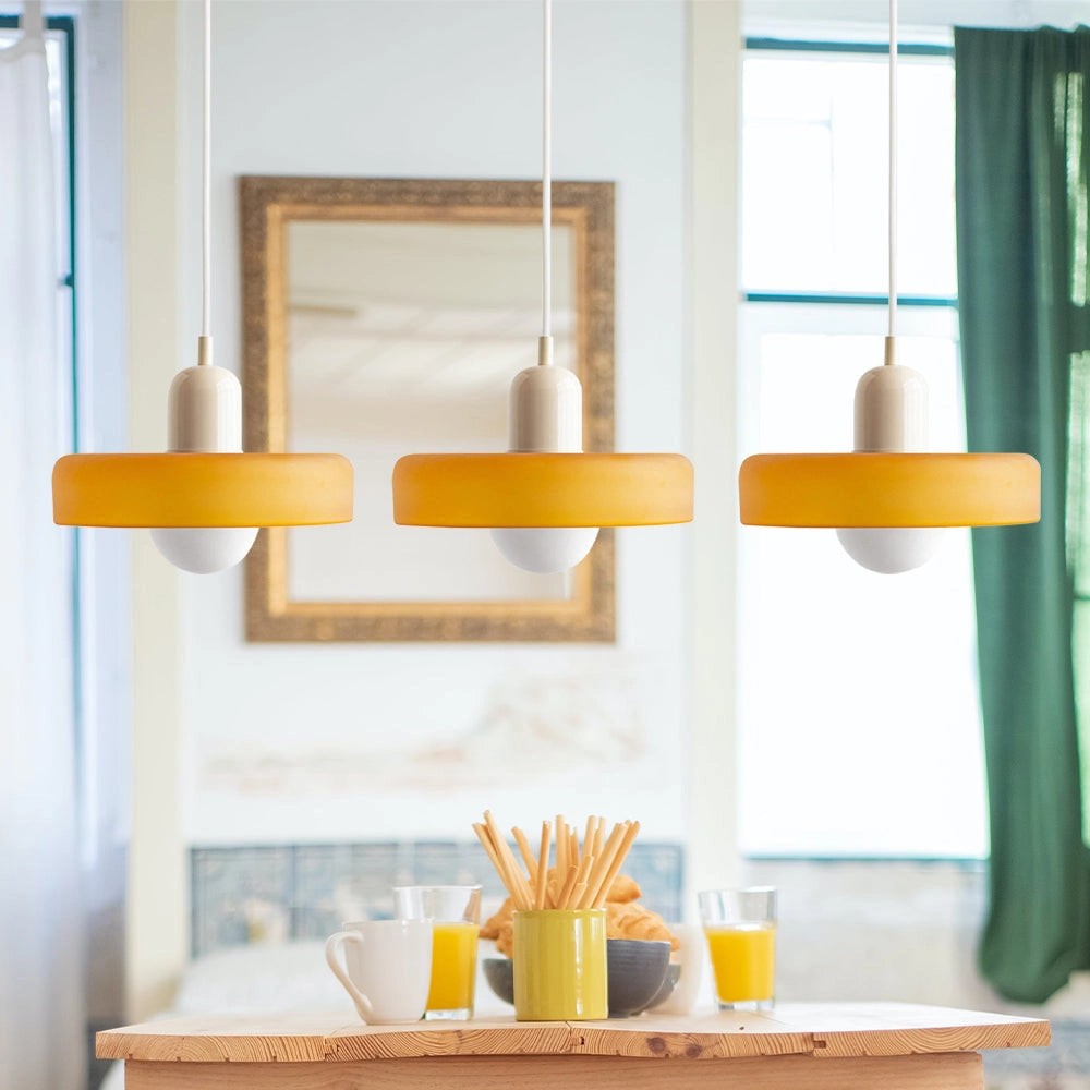 Modern brass pendant lights with three heads, orange and white color scheme, red cable, hanging in an industrial-style kitchen with wooden shelving and rectangular windows in the background.