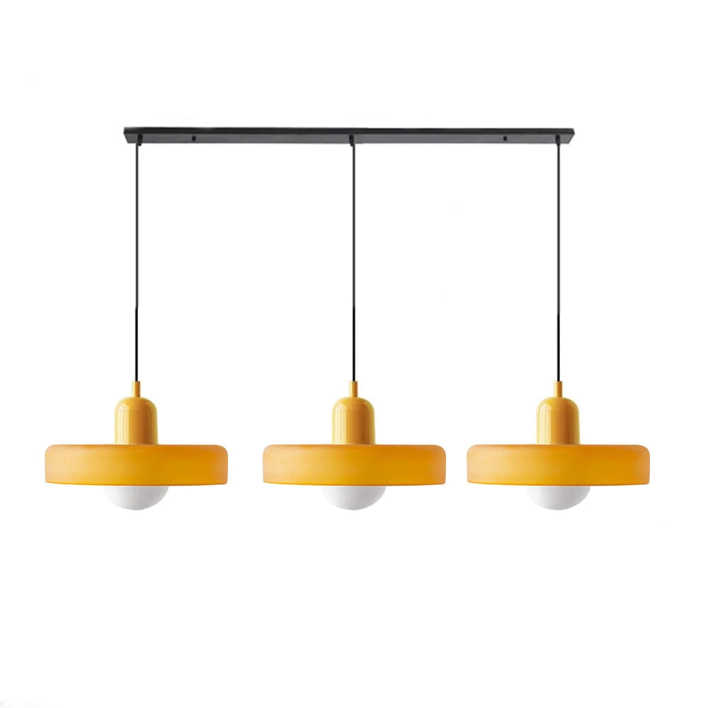 Modern brass pendant lights with three heads, adjustable height, featuring red cables, suspended above a wooden table, with plants and shelving in the background.