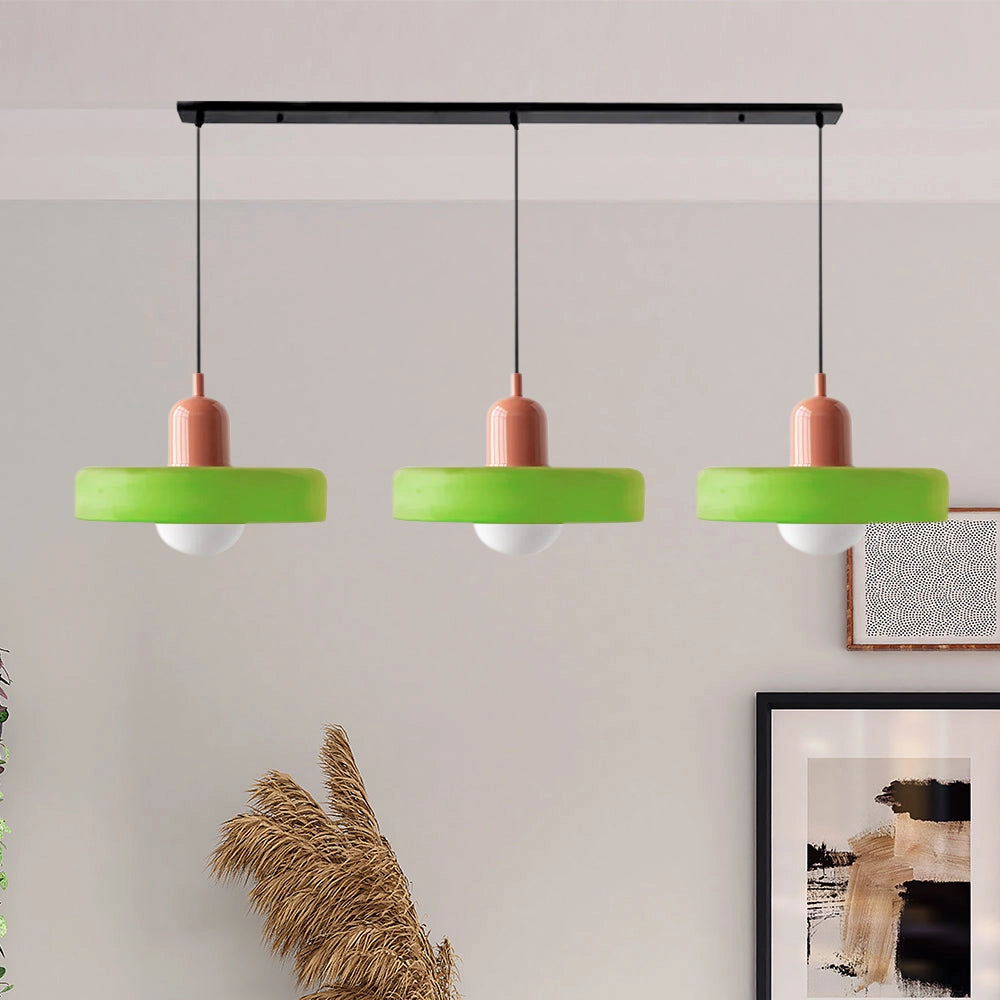 Modern brass pendant lights with three adjustable heads, featuring a matt green and pink finish, suspended by a red cable, hanging above wooden shelving in an industrial kitchen setting.