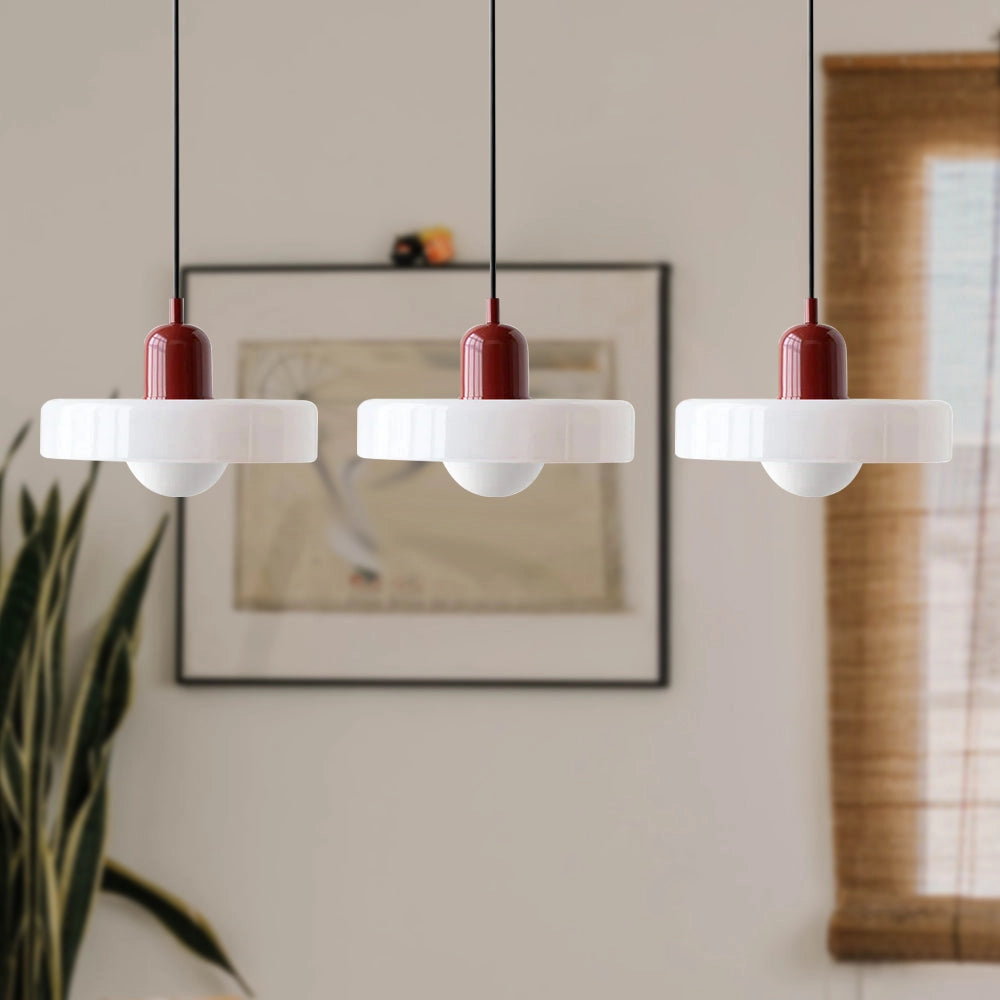 Modern brass pendant lights with three adjustable heads, featuring white shades and red cables, hanging in an industrial kitchen setting with wooden shelving, houseplants, and hardwood flooring.