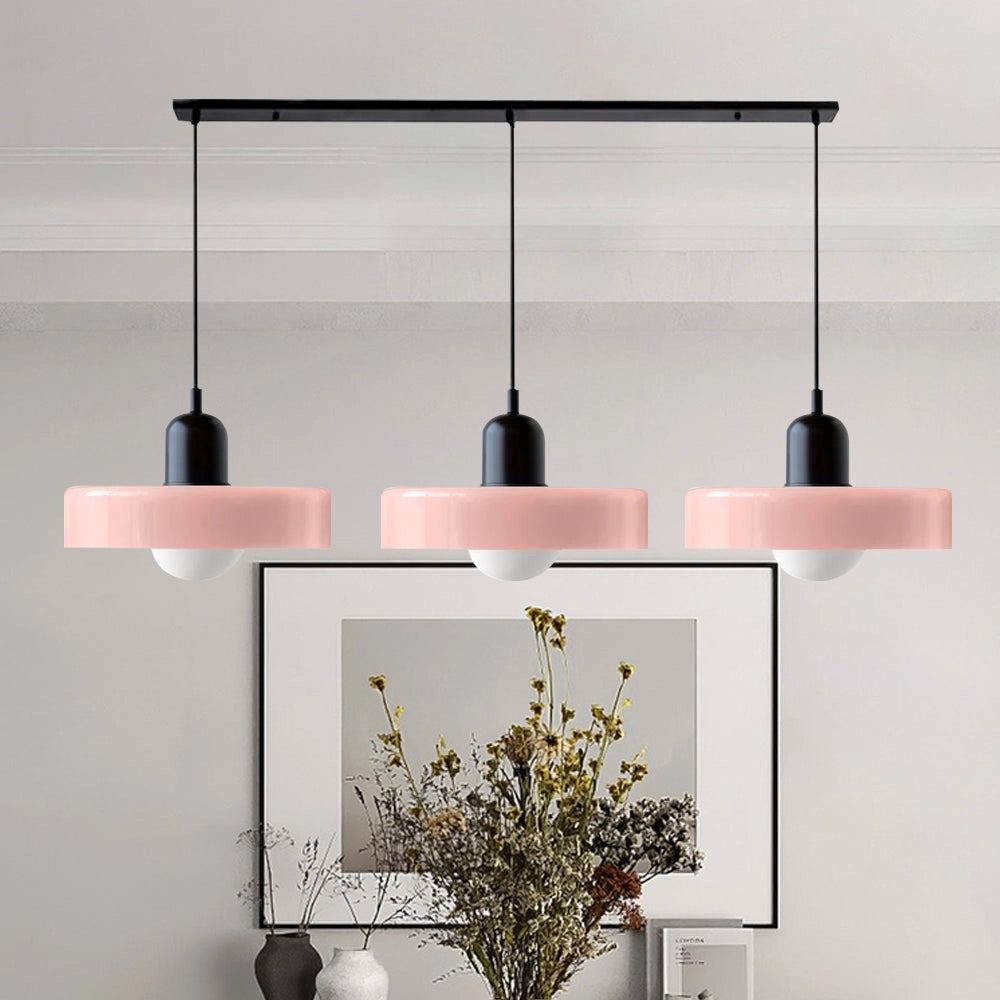 Modern brass pendant lights with three adjustable heads and red cables, styled in an industrial kitchen setting with pink and black accents, surrounded by houseplants and various decorative items on wooden shelves.