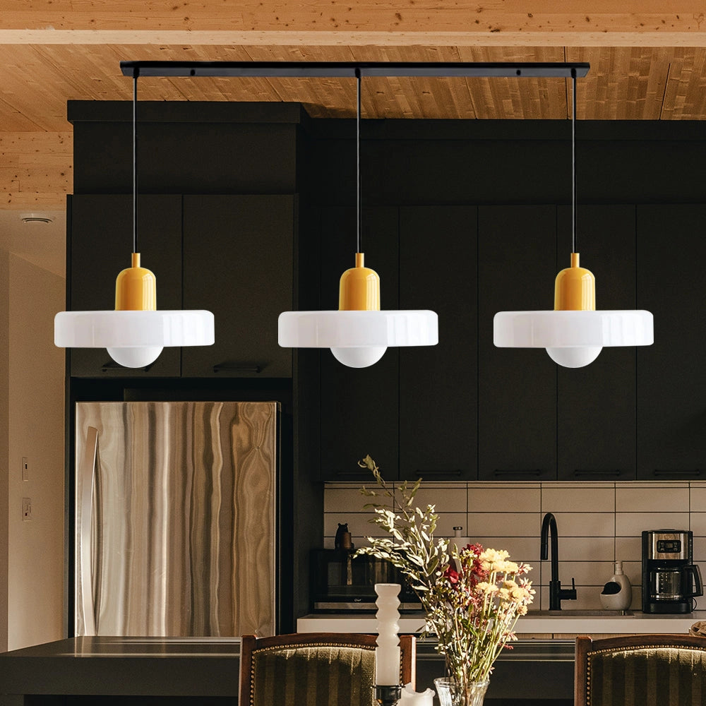 Modern brass pendant lights with adjustable red cable, featuring three white and yellow heads, hanging in an industrial kitchen setting with wooden elements and houseplants visible.