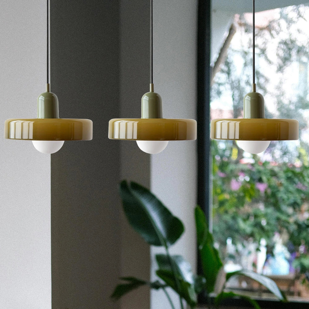 Modern brass pendant lights with adjustable red cables and three green heads, hanging above an industrial kitchen setting with wooden shelving and glass candle holders.