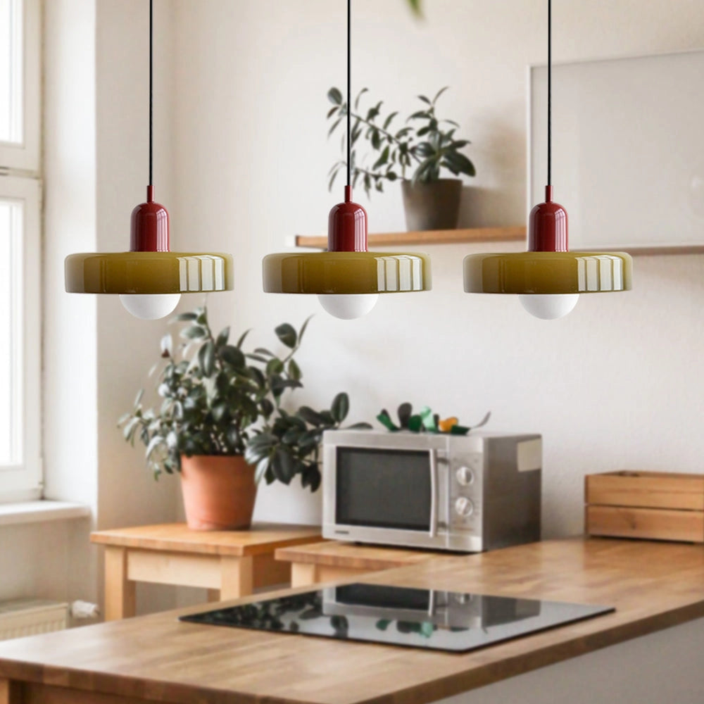 Modern brass pendant lights with red cables, adjustable, featuring three green and red heads, hanging above an industrial kitchen table with houseplants and wooden shelving in the background.