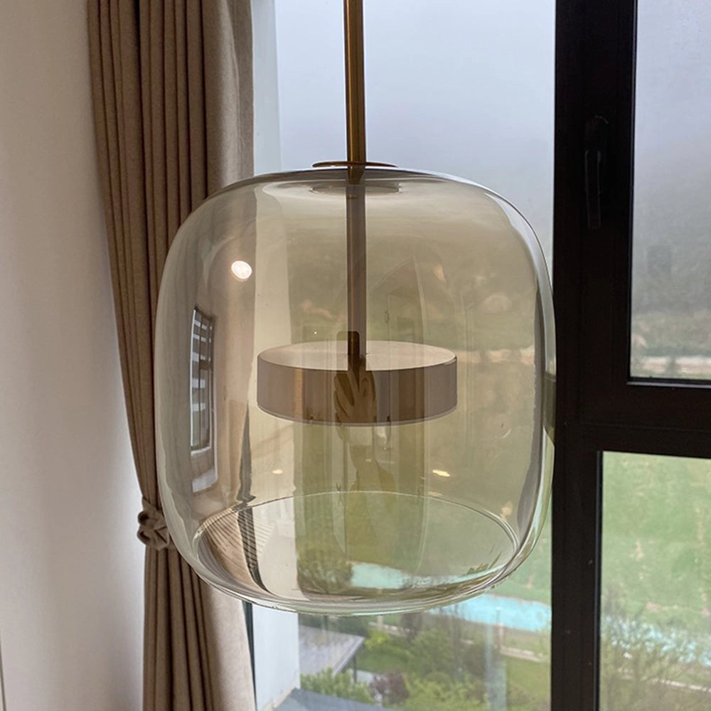 Modern brushed brass glass pendant light with a rectangular shape, featuring a transparent glass shade, hanging in a kitchen or dining area with wooden accents and window treatments in the background.