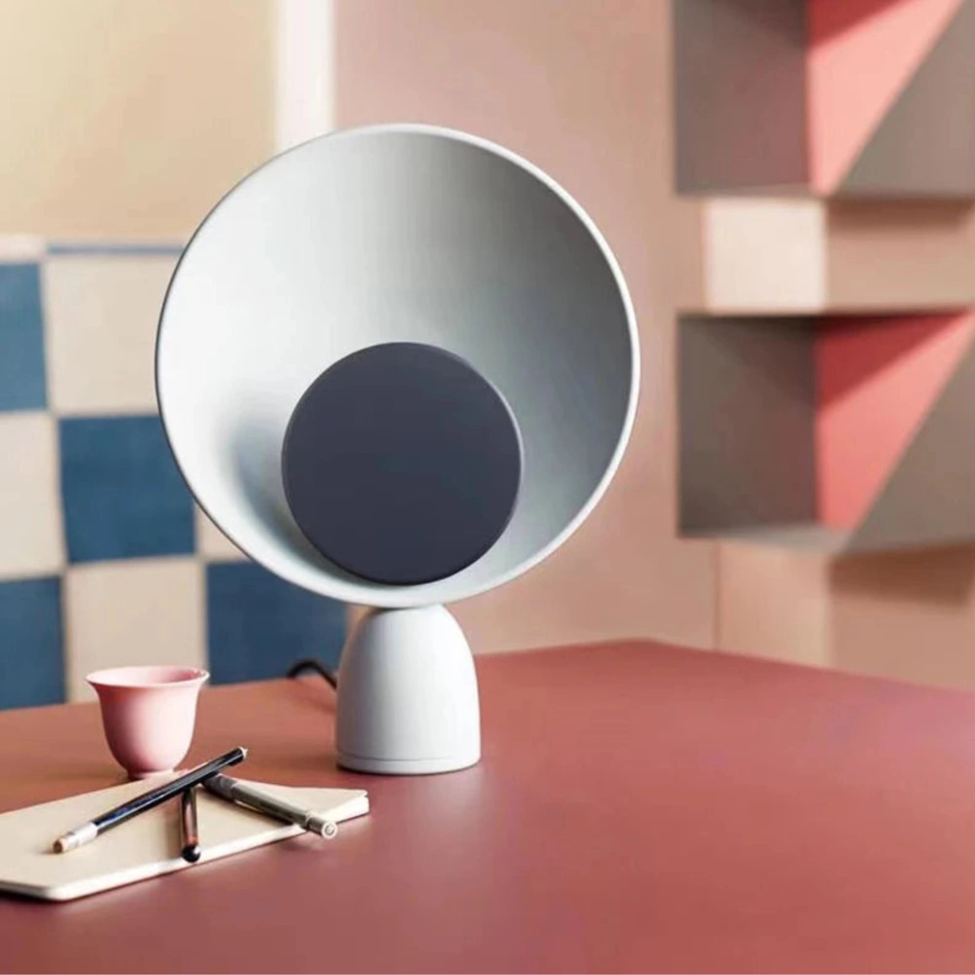A modern circular acoustic speaker with a sleek design, placed on a wooden table near a window, surrounded by various tableware items including cups and dishes.