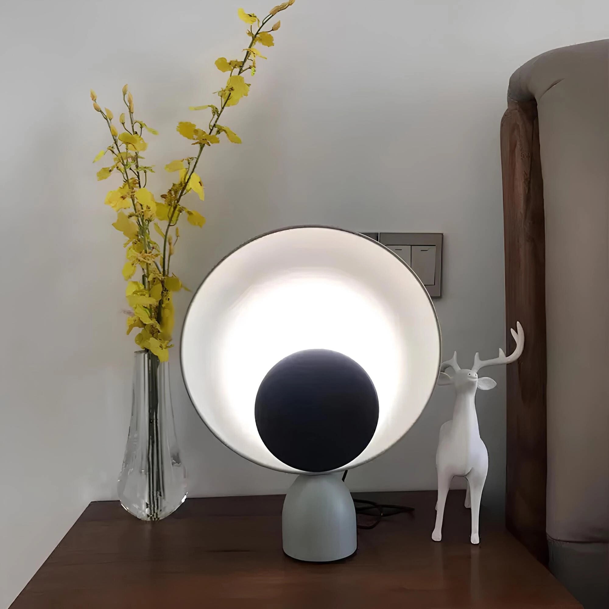 Modern circular acoustic speaker in black and cold white color placed on a wooden table in a stylish interior setting with a grey couch, indoor plant, and contemporary lighting.