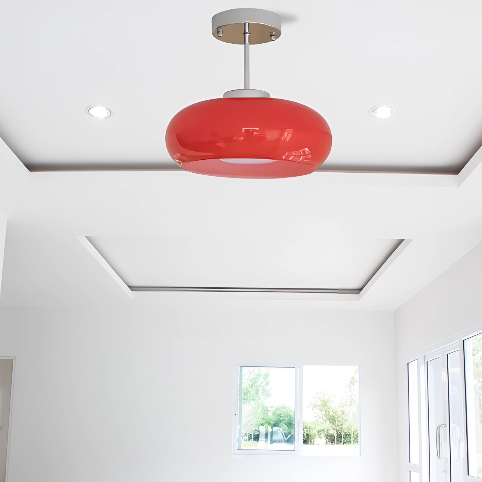 Modern circular LED pendant light with a minimalist red shade, suspended from a white ceiling, providing ambient lighting in a contemporary setting featuring large windows and potted plants.