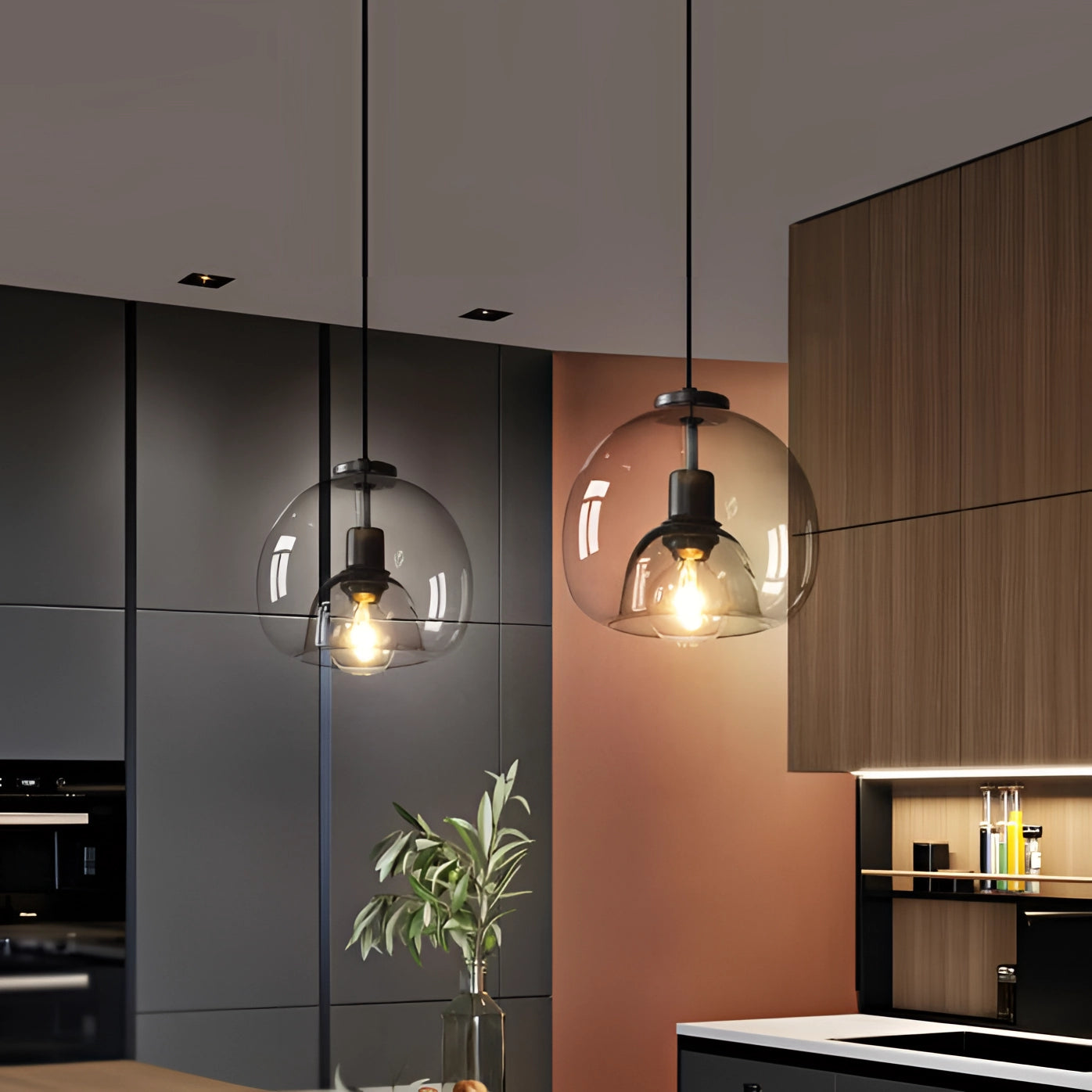 Modern clear glass pendant lights in a stylish globe design hanging in an interior setting, with houseplants and wooden cabinetry in the background.