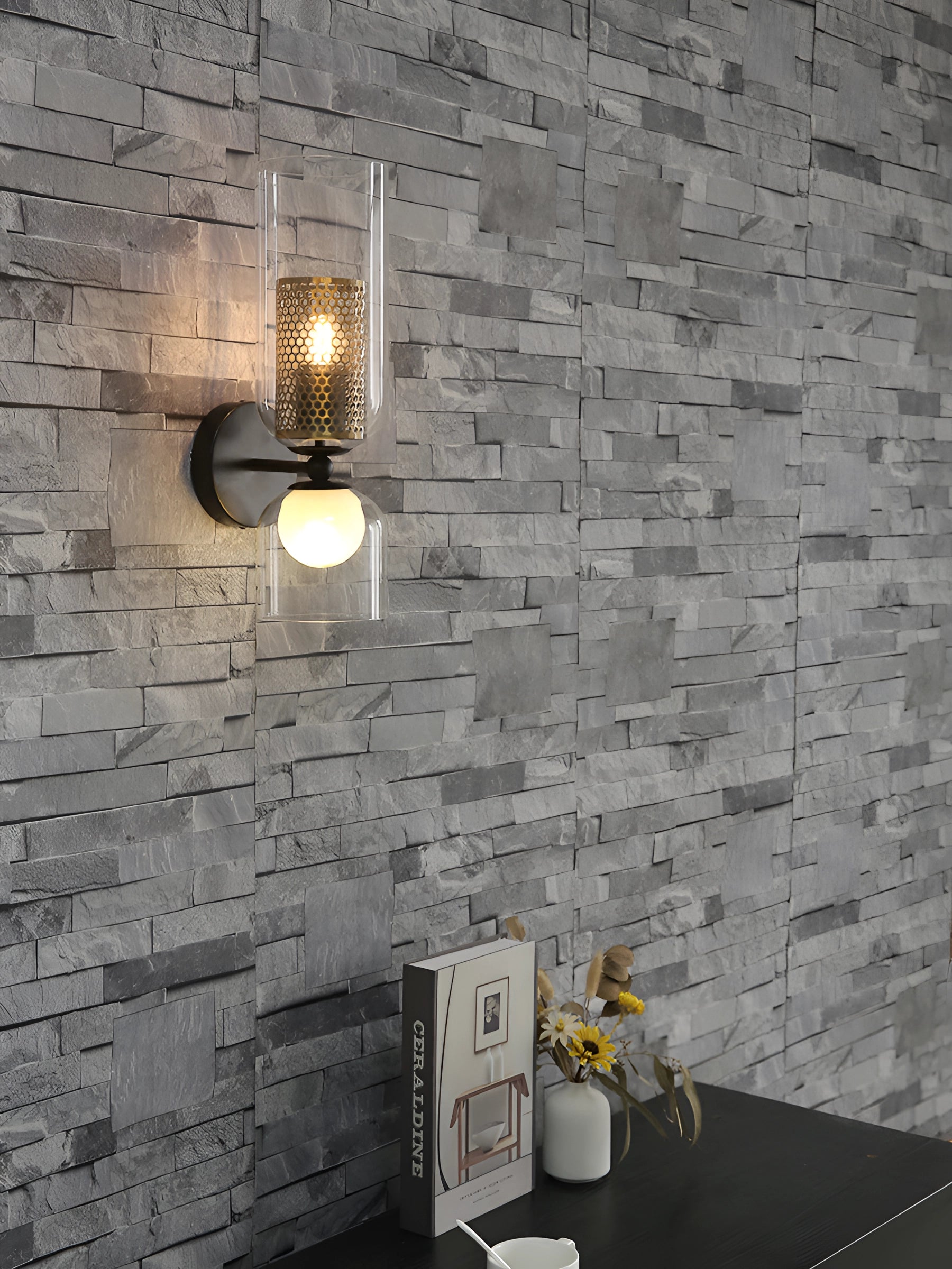 Modern cylindrical glass wall sconce with a bronze mesh shade mounted on a wooden wall, surrounded by brick accents and houseplants.