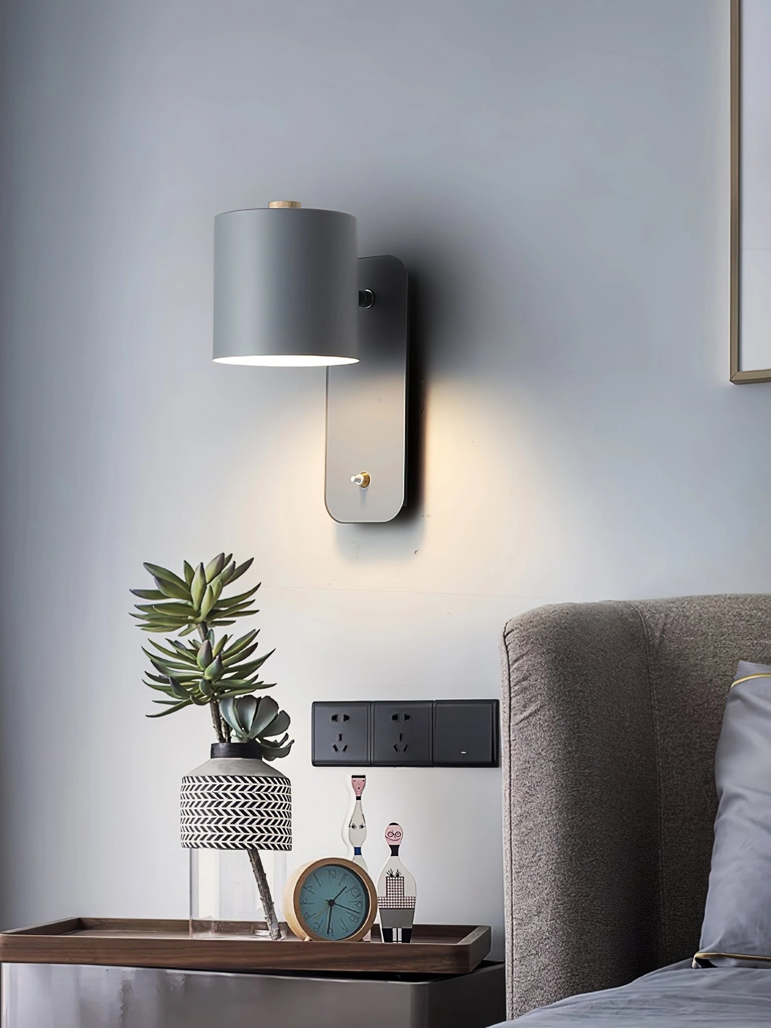 Modern cylindrical LED wall lamp in a minimalist metal sconce design installed on a grey wall, surrounded by contemporary interior decor including wooden elements and a houseplant.