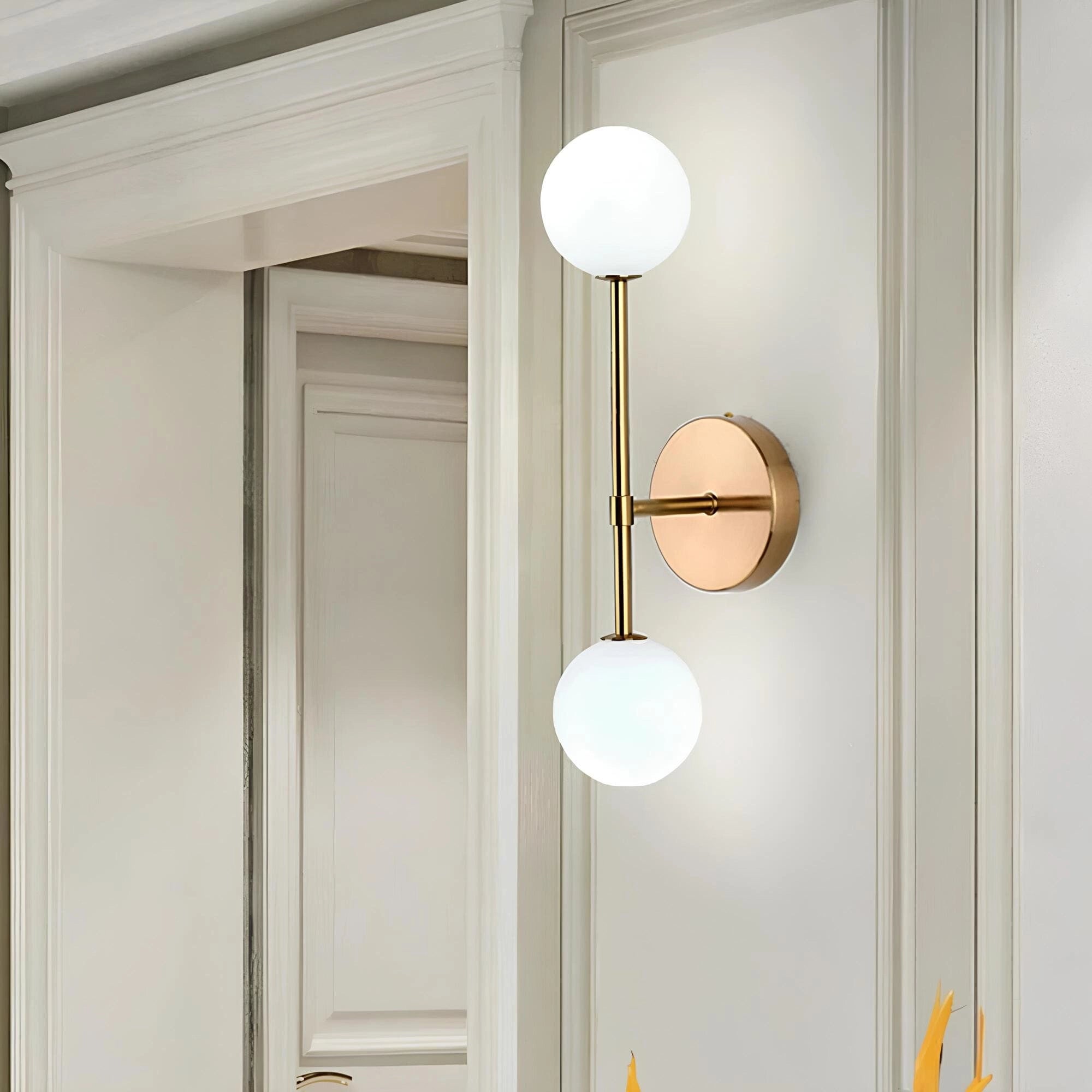 Modern double globe wall sconce in gold finish, featuring minimalist LED lighting, mounted on a wood-paneled wall next to a door.