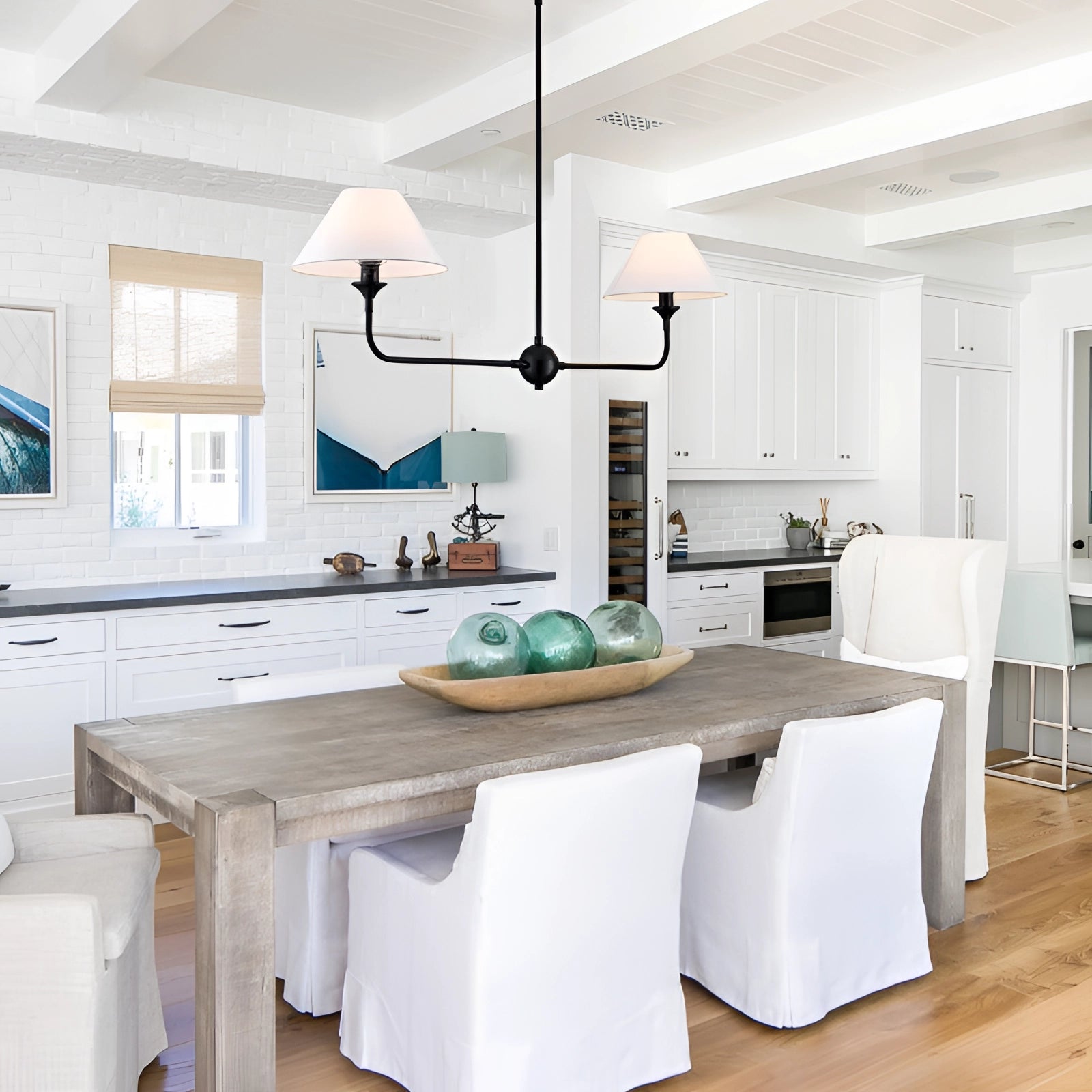 Modern dual lamp black LED chandelier with double cone pendant light in a white variant, hanging above a stylish kitchen interior featuring wooden cabinetry, a grey countertop, and a well-furnished dining area with a table and flooring.