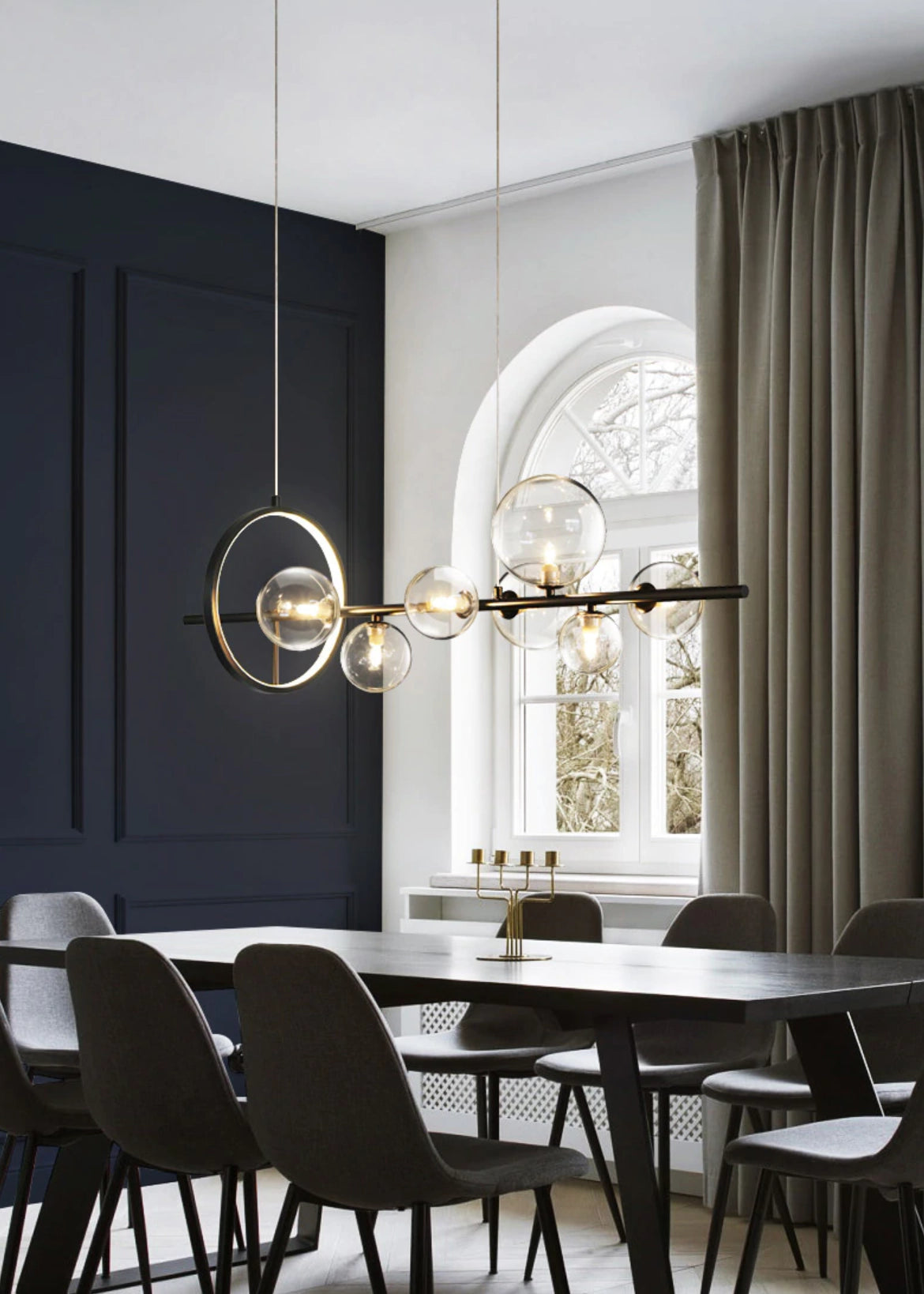 Modern geometric brass and glass chandelier hanging above a wooden table in a stylishly decorated interior with large windows and indoor plants.
