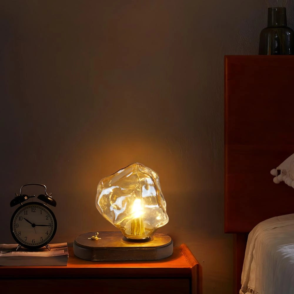 Modern geometric glass desk lamp with wooden base, emitting warm amber light, placed on a table next to an alarm clock.