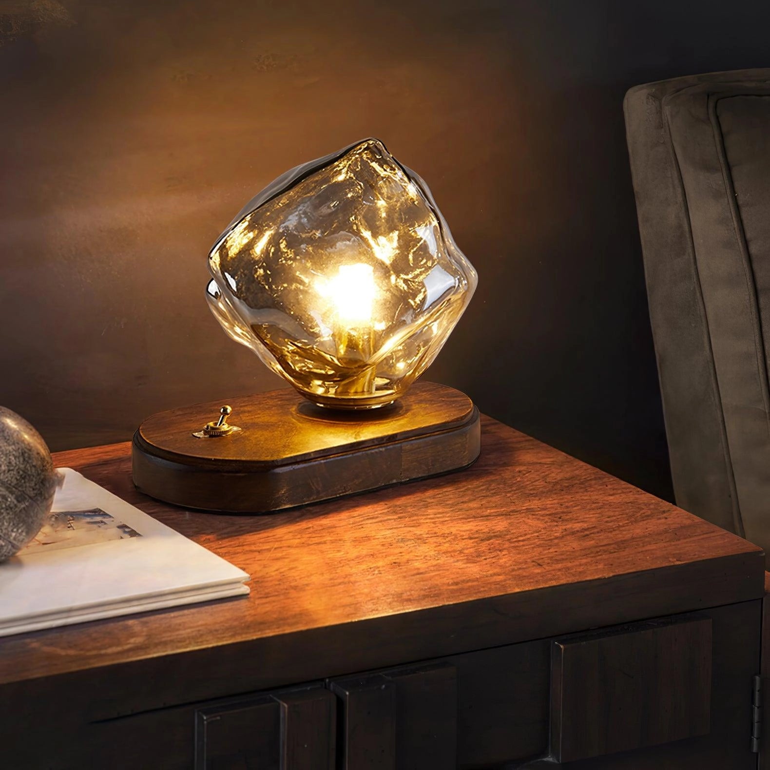 Modern geometric glass desk lamp with amber light, featuring a wooden base, positioned on a table with surrounding wooden cabinetry.