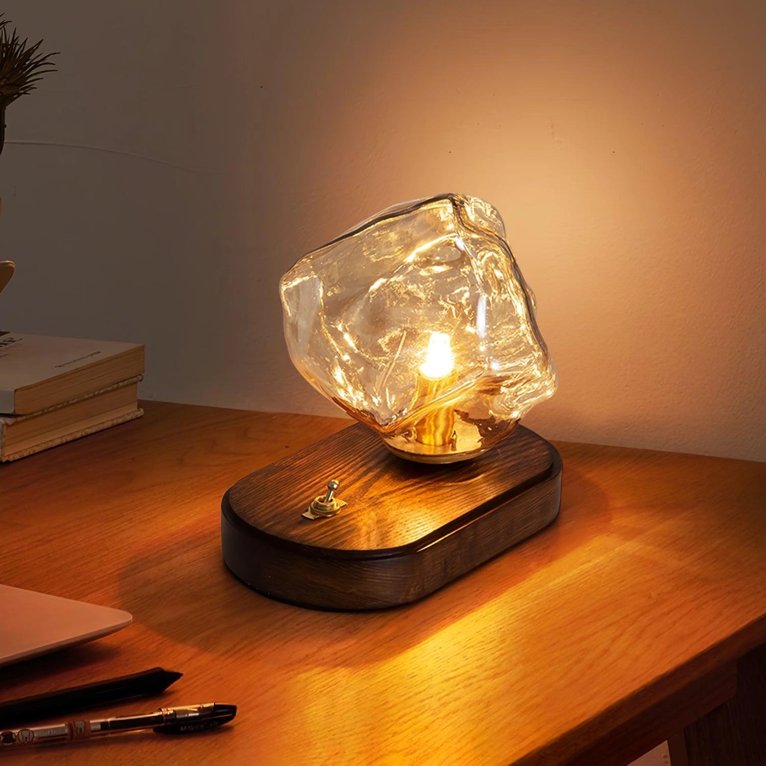 Modern geometric glass desk lamp with amber glow, wooden base, and rectangular shape, placed on a desk next to a houseplant in a flowerpot.
