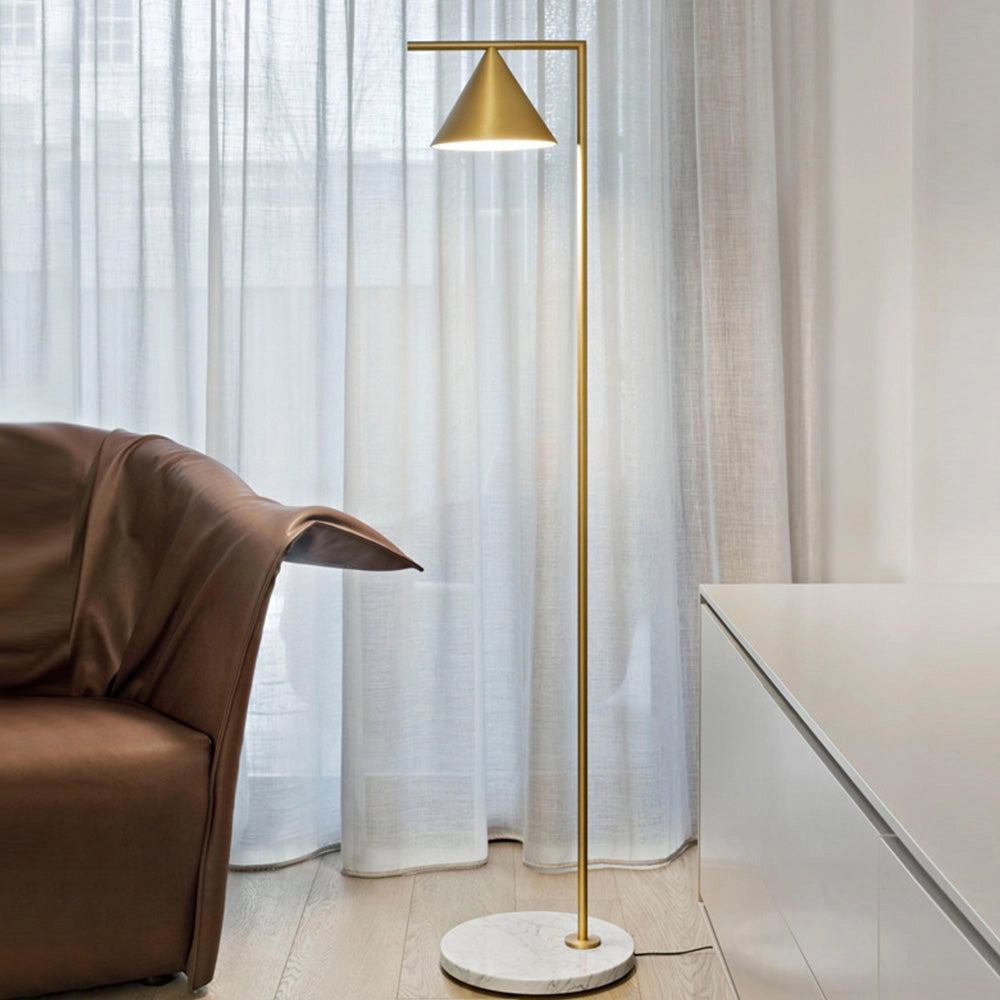 Modern geometric gold floor lamp with a minimalist design, featuring a warm white light. The lamp is placed in a stylish interior setting, complementing the wooden floor and neutral curtains in the background.