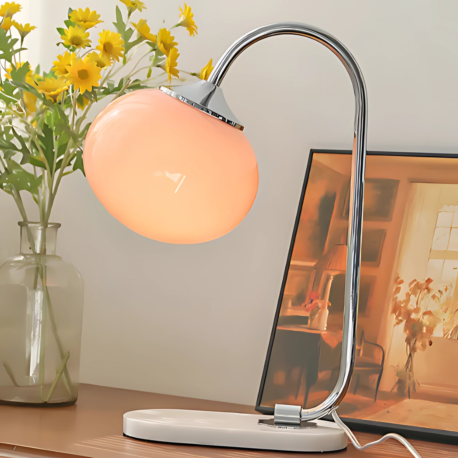 A modern table lamp with a minimalist curved design, featuring a gold finish with silver pink accents, placed on a desk near a computer and flowers.