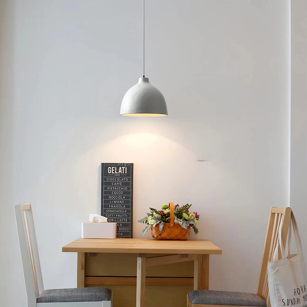 Modern industrial concrete pendant light with a Scandinavian yellow accent, featuring a grey finish and an 11-inch diameter, hanging above a wooden table in a stylishly designed interior with plants, shelving, and light grey walls.
