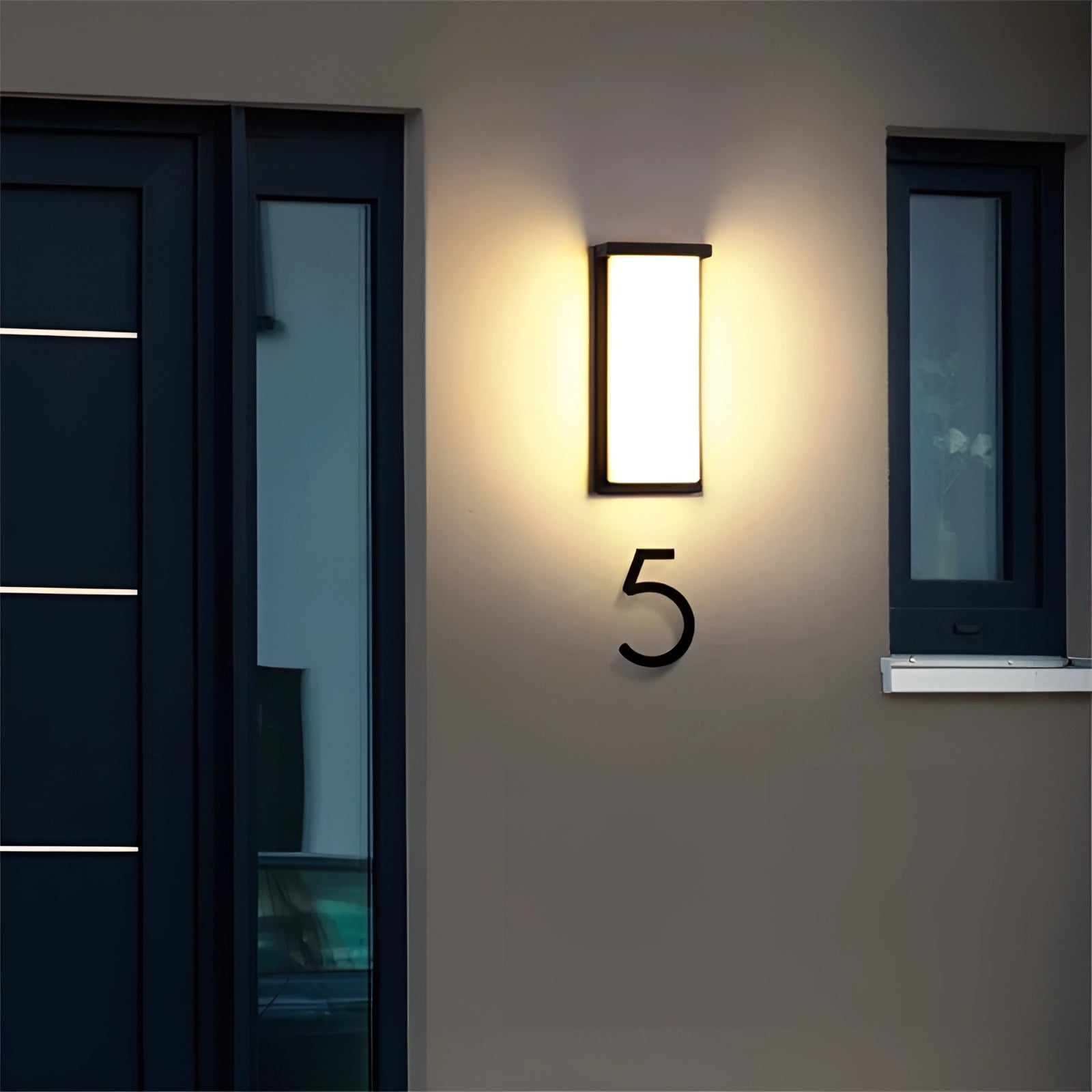 Modern LED outdoor wall light fixture mounted vertically on a wooden exterior wall next to a door with a glass panel and metal handle. The rectangular design features clean lines and contemporary styling, suitable for enhancing outdoor spaces.
