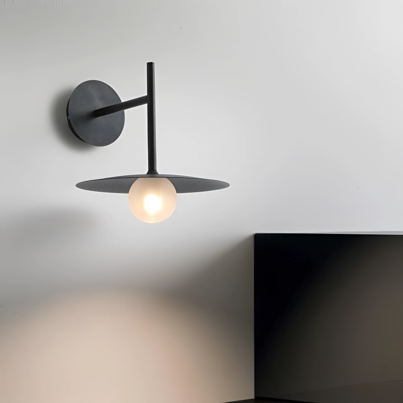 Modern matte black minimalist wall sconce with circular design, featuring a sleek light fixture mounted on a wooden base, casting soft shadows, ideal for bedroom and hallway lighting.