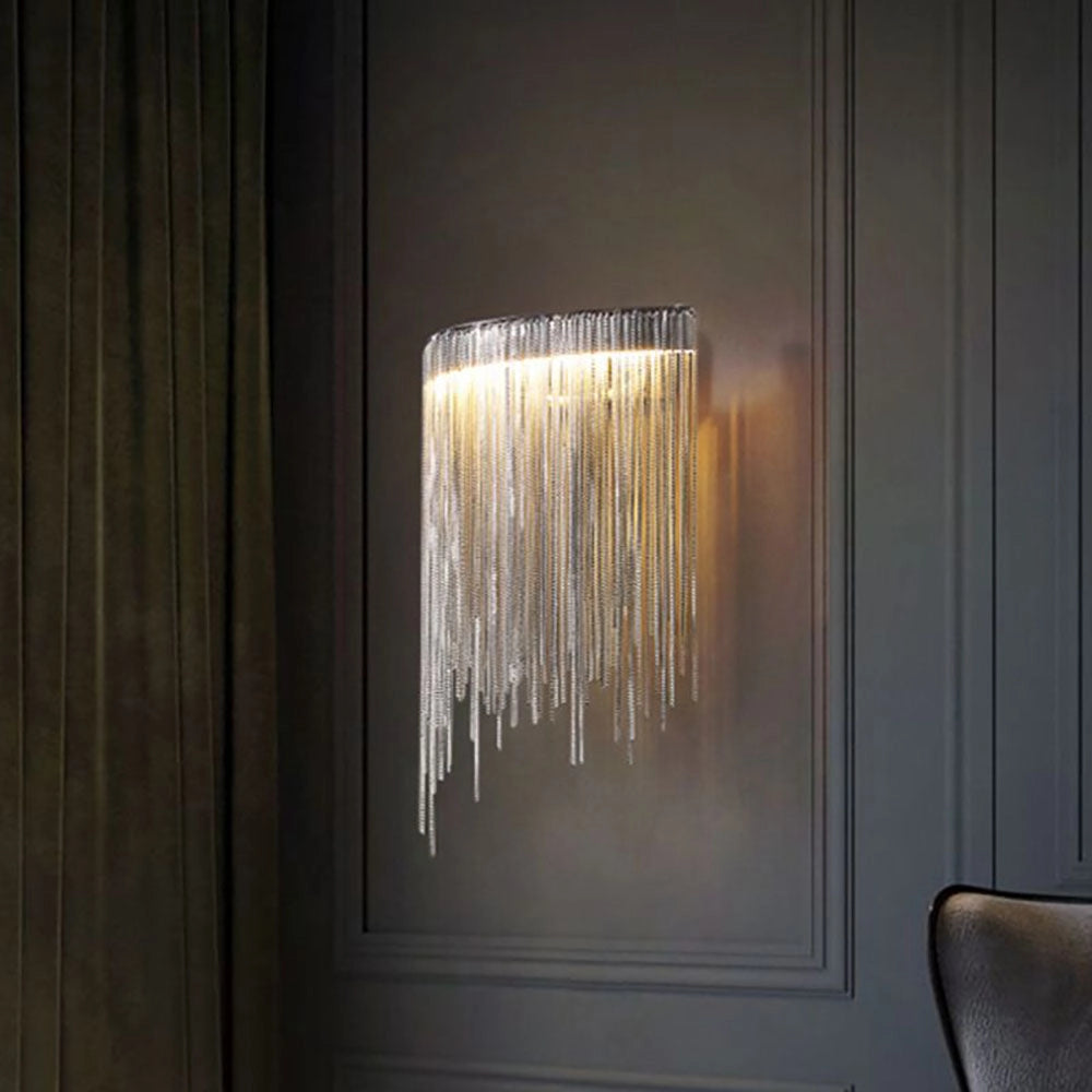 Modern silver metallic fringe chandelier hanging from the ceiling, with a luxurious contemporary design, featuring reflective glass and metal elements. The background showcases a hardwood floor and a wooden art piece.