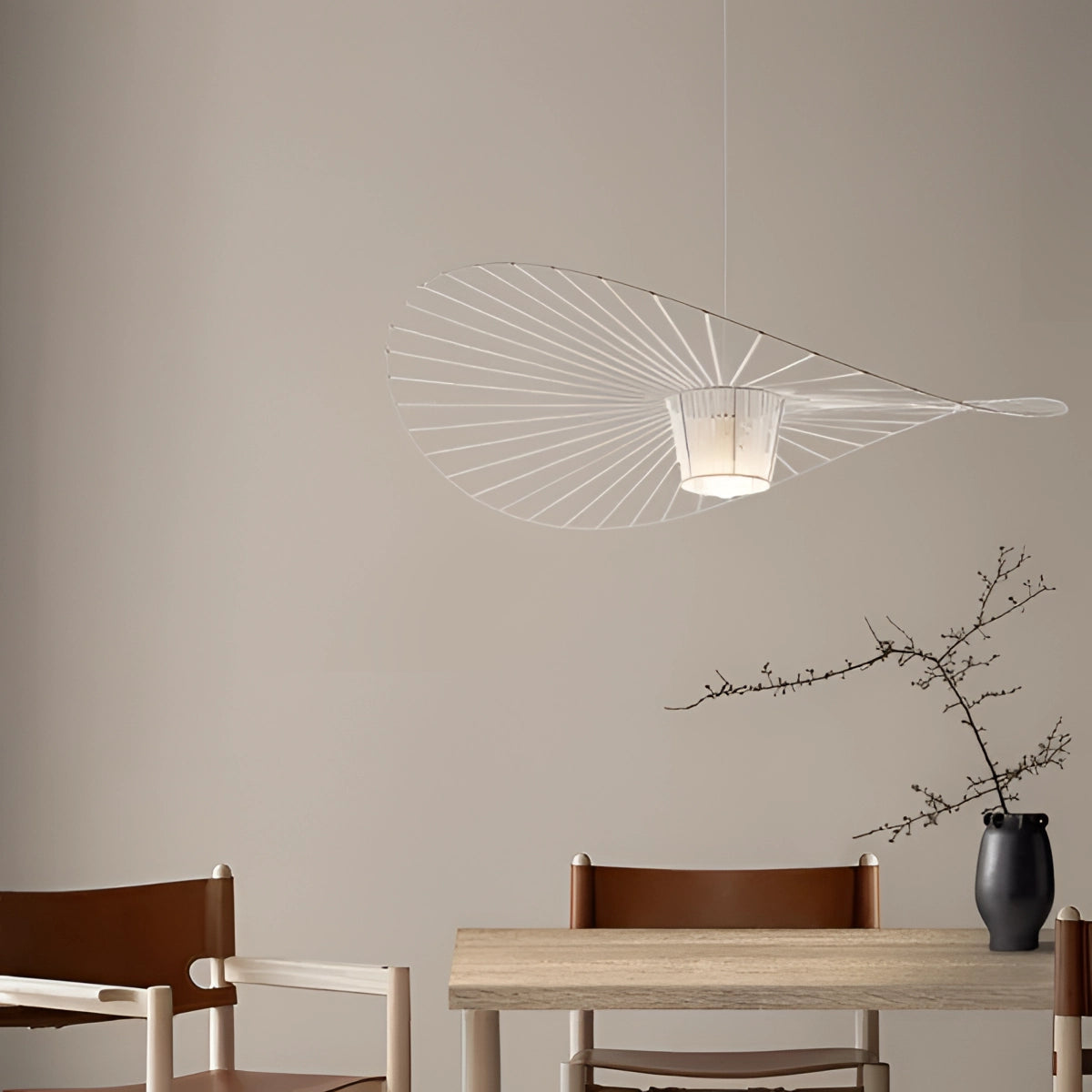 A modern minimalist ceiling fan with sleek, silent wing-shaped blades is installed on a white ceiling in a stylish interior space. The room features contemporary furniture, including a wooden table and chairs, with art pieces and plants enhancing the decor. The overall design exudes a sense of elegance and simplicity.