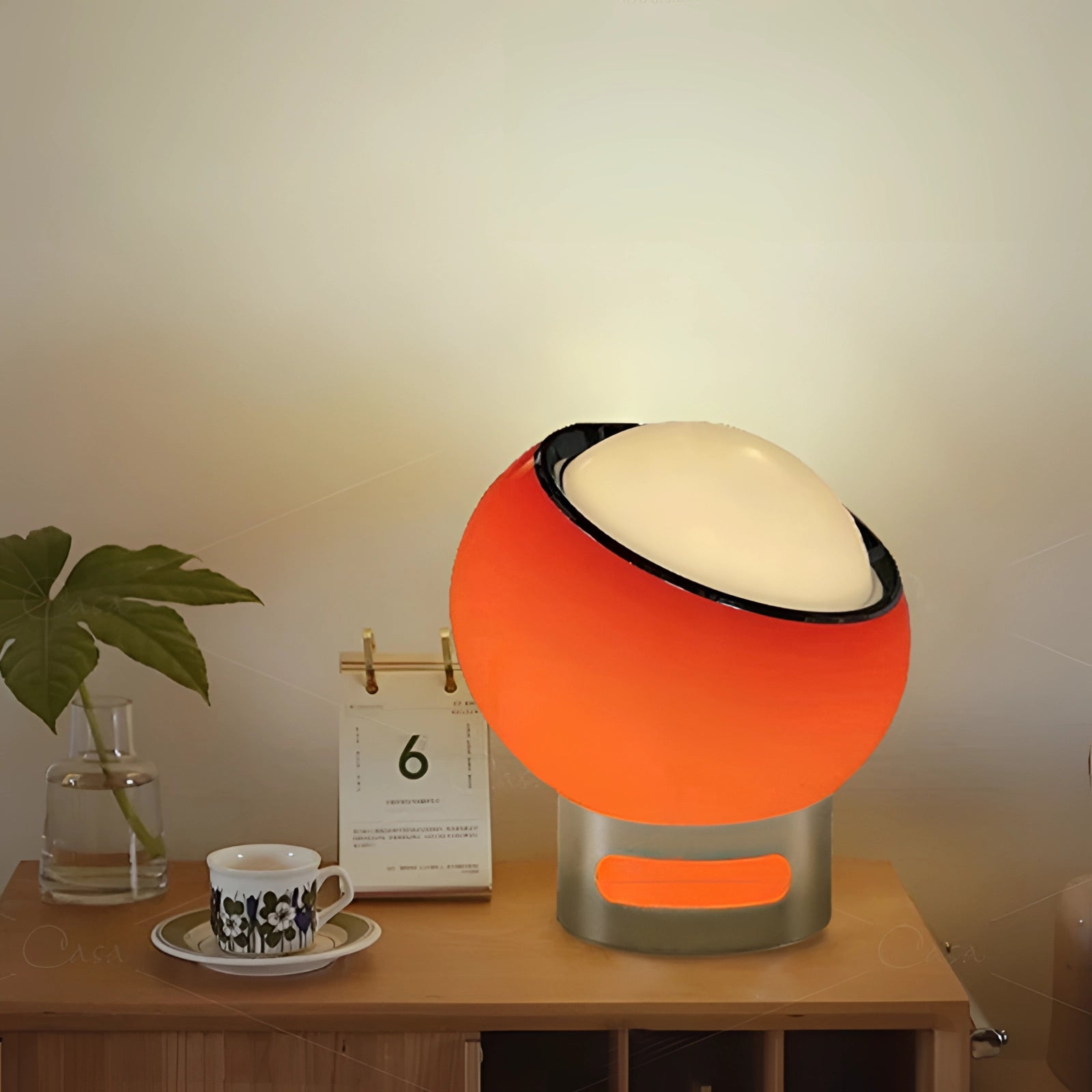 Modern minimalist circular LED bedside lamp with sleek metallic base placed on a wooden table, next to a small potted plant in a stylish interior setting.