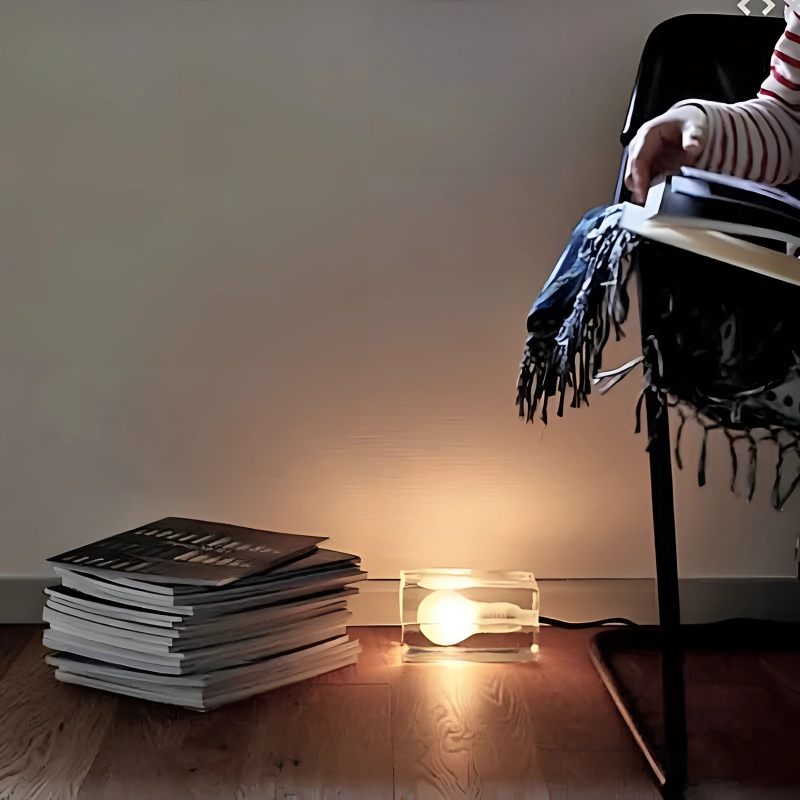 A modern minimalist clear glass cube table lamp with a wooden base, emitting a soft glow in a dimly lit room, set on a hardwood floor.